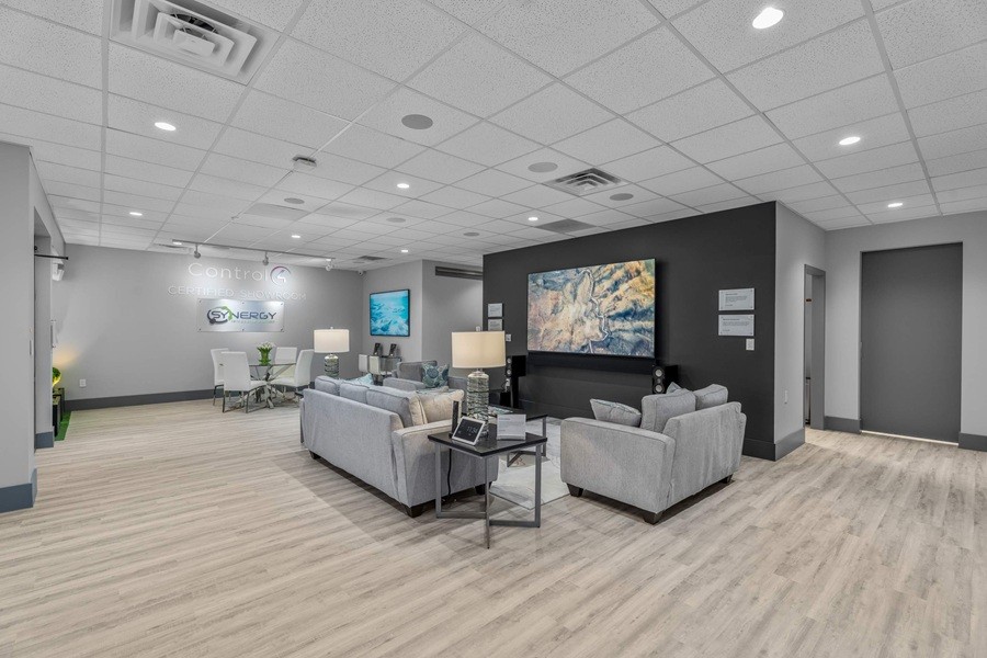 Synergy Integrated Systems showroom featuring a seating area with two sofas, wall-mounted display, smart tablet on side table, and Control4 Certified signage.