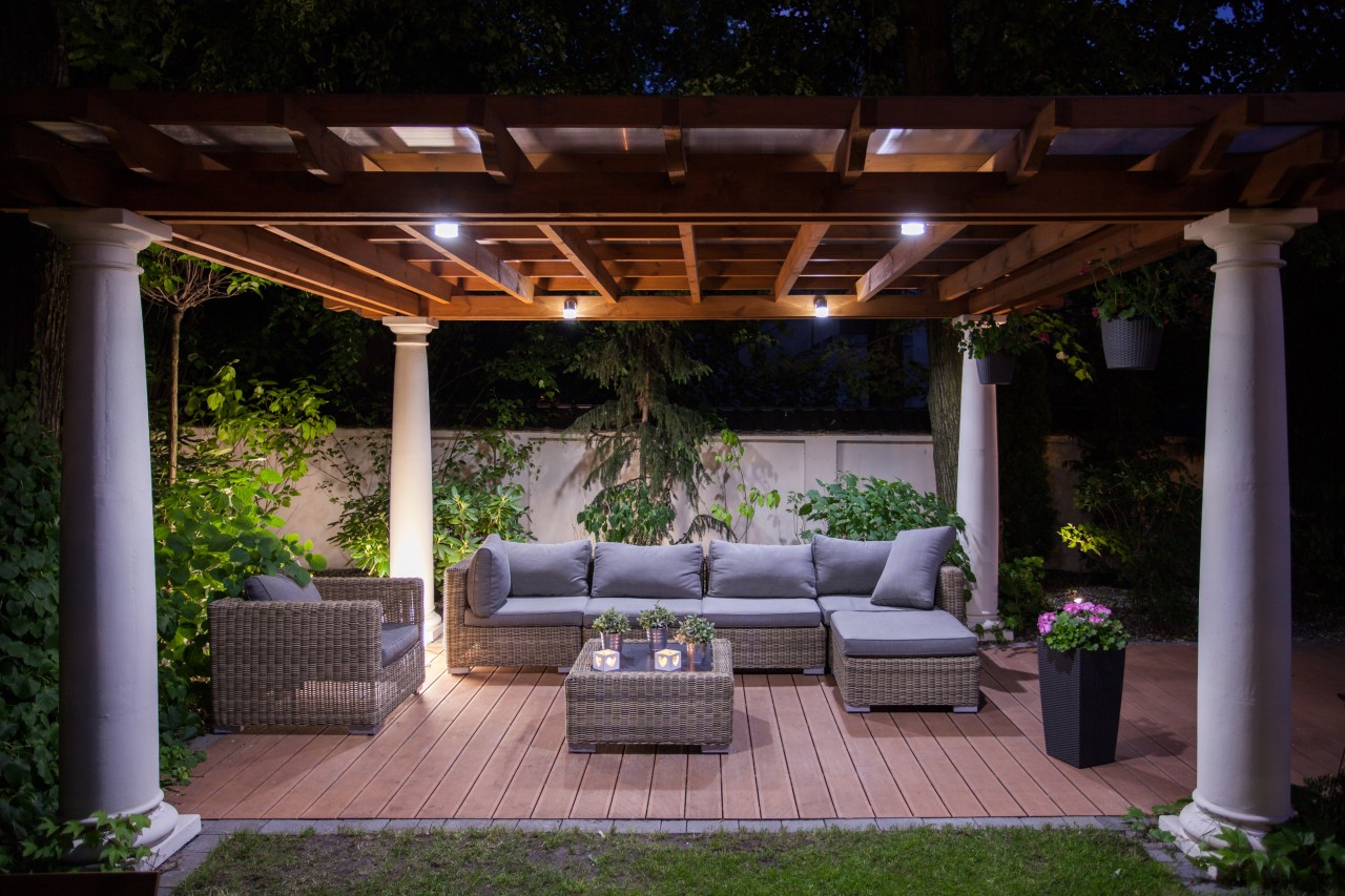 A covered patio with couches, plants, and an end table at night time