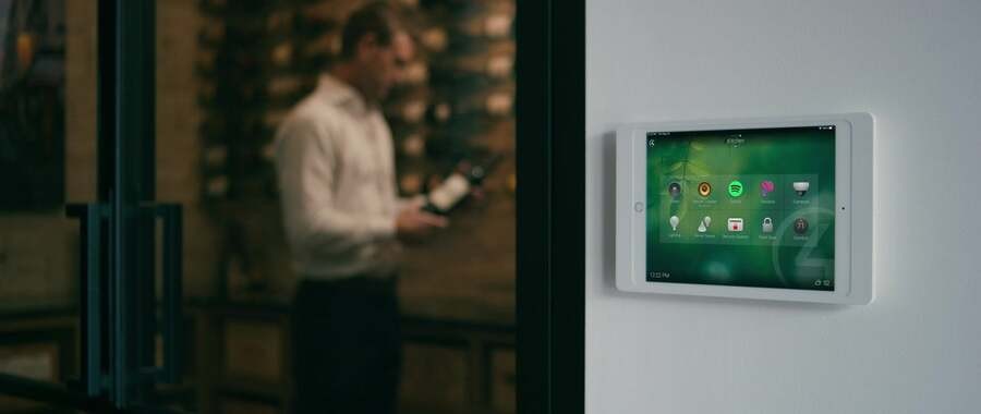 A wall with a keypad displaying a Control4 system while a man chooses a bottle of wine in the back. 