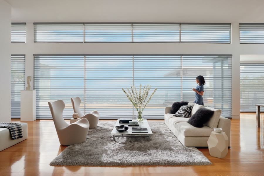 A woman in a living room looking out picture windows covered by sheer blinds.