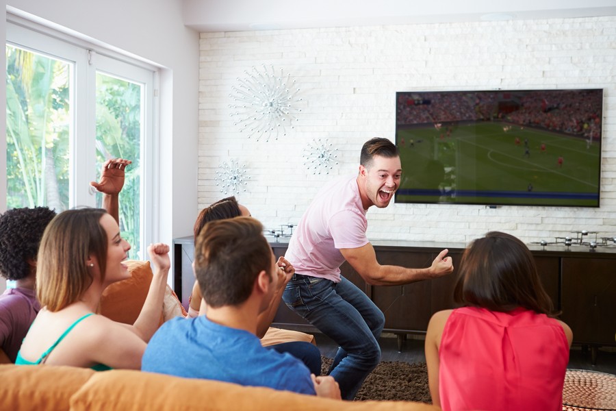  Image is of people on a couch in a media room in a modern smart home, cheering at a sports game on the TV.